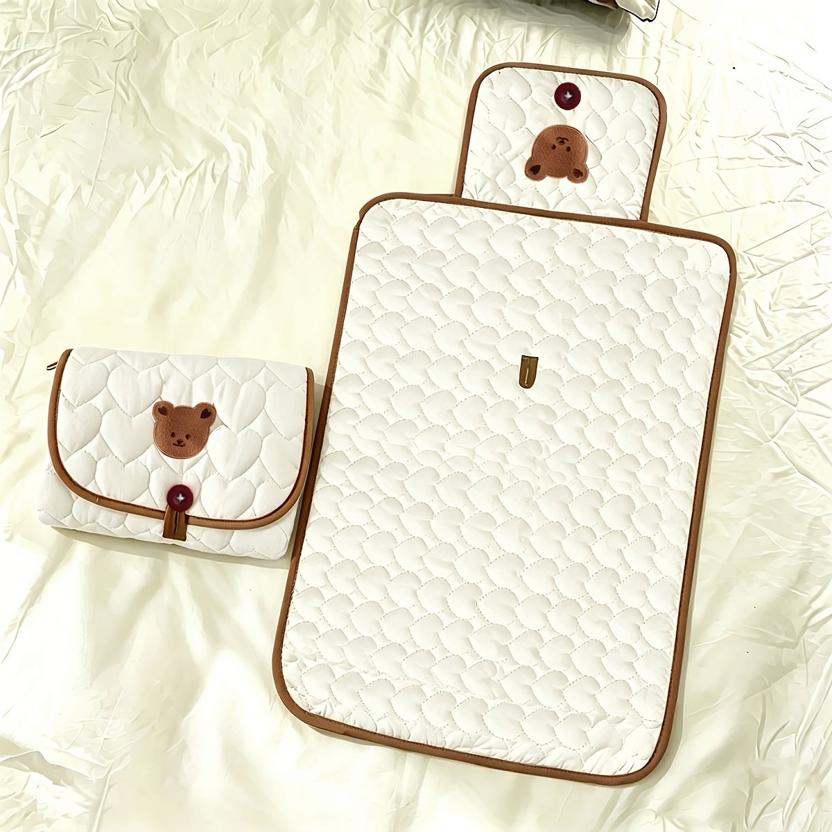 A quilted white baby blanket set with brown trim, featuring a large rectangular blanket and a matching small pouch, both decorated with a brown bear face applique and a red button.