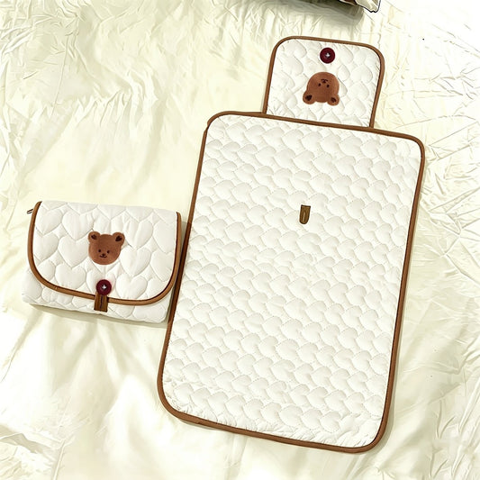 A quilted white baby blanket set with brown trim, featuring a large rectangular blanket and a matching small pouch, both decorated with a brown bear face applique and a red button.