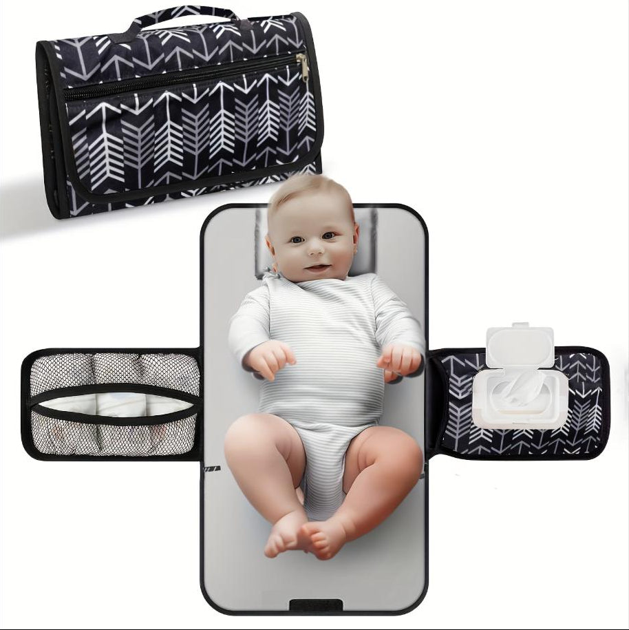 Portable baby diaper changing pad with black arrow print, unfolded flat showing baby lying on cushioned mat with mesh storage pocket and wipes compartment